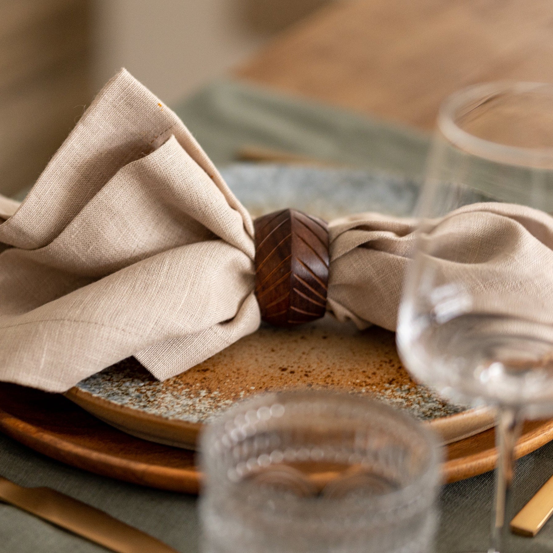 Neatly folded beige napkin on a wooden plate with glasses and cutlery in the background.