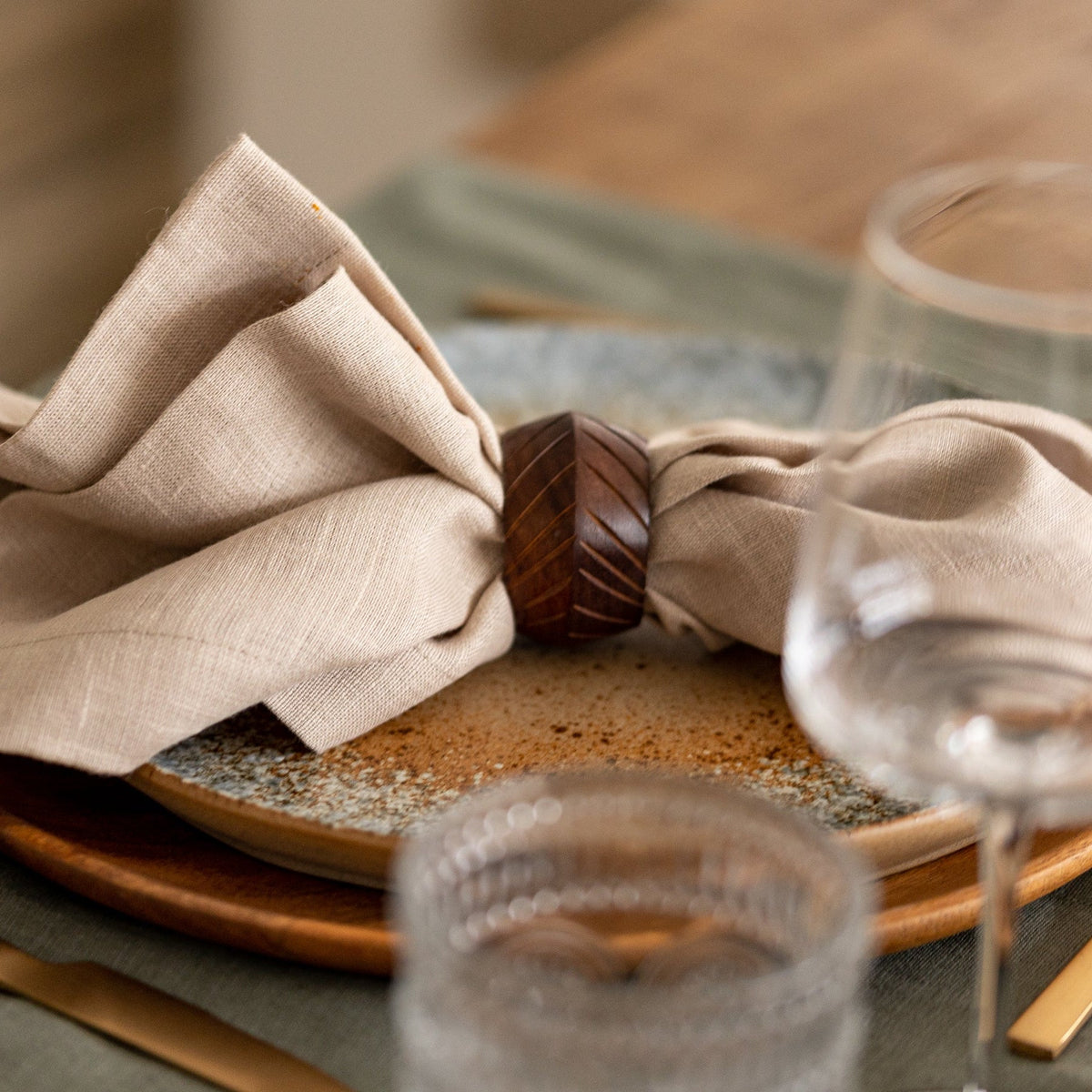 Neatly folded beige napkin on a wooden plate with glasses and cutlery in the background.